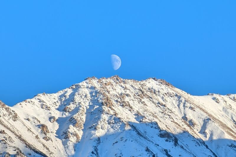 A snow-capped mountain peak stands against a clear blue sky, with a half-moon visible just above the peak.