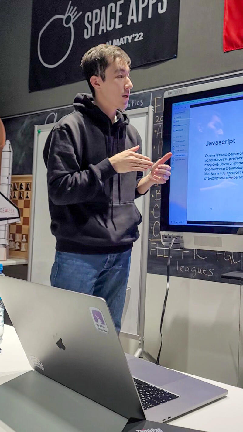 A man in a black hoodie and jeans is presenting in front of a screen displaying text about Javascript, with a laptop in the foreground and a 'Space Apps Almaty' banner in the background.