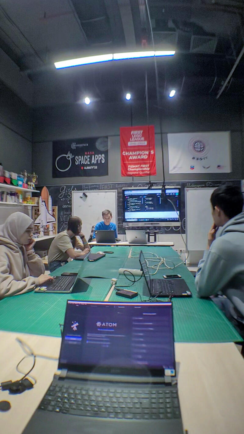 A group of people are sitting around a table with laptops, working on a project, with '#ATOM' visible on one of the laptop screens and banners for NASA Space Apps and FIRST LEGO League in the background.
