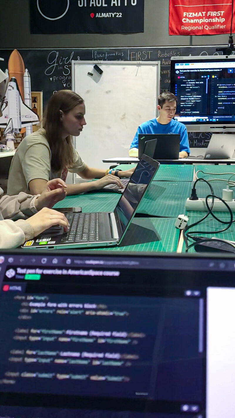 The image shows a group of people working on laptops at a table, likely coding or programming, with banners for 'Space Almaty' and 'Fizmat First Championship' in the background, along with a whiteboard and a display showing lines of code.
