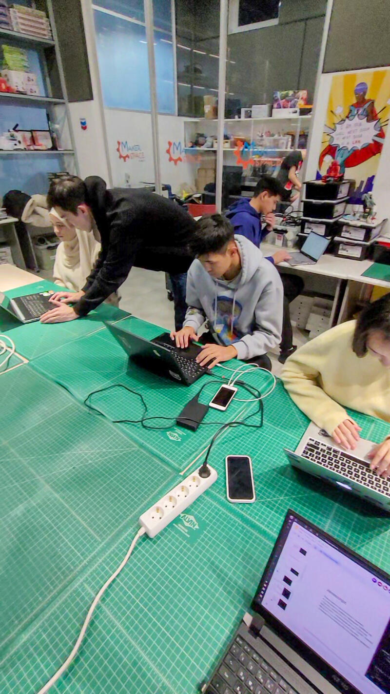 Several people are sitting at a green gridded table, working on laptops and charging their phones using a power strip.
