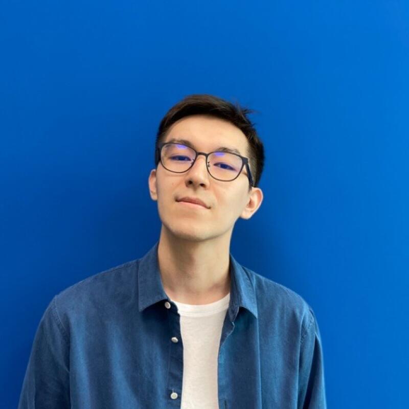 A man with glasses and a blue denim shirt stands against a blue background.