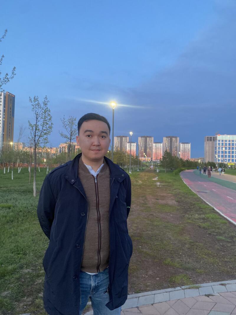 A man in a blue jacket and jeans stands in a park with apartment buildings and a blue sky in the background.