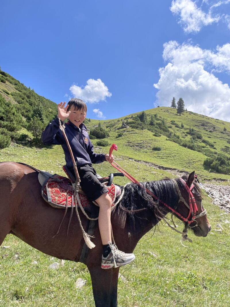 A young boy is riding a horse in a grassy field, waving at the camera with a mountain and blue sky in the background.