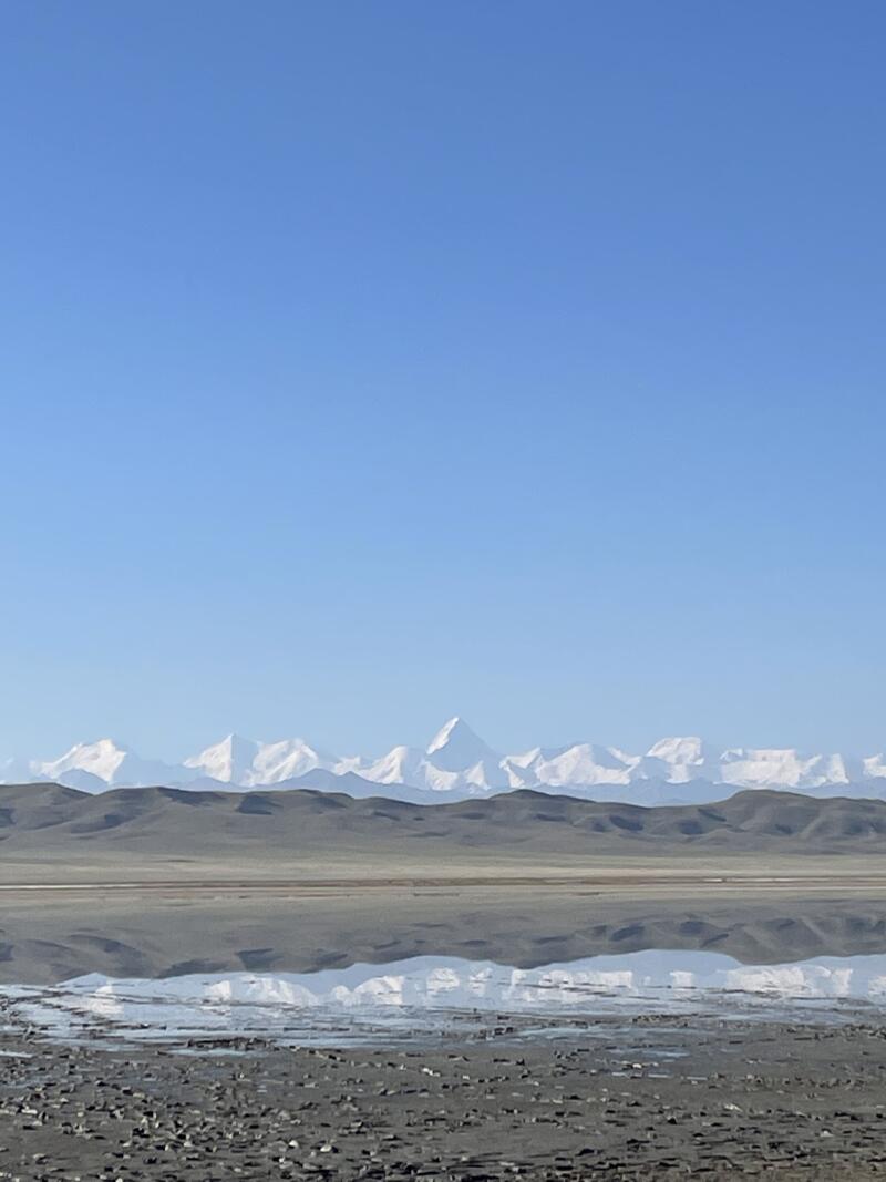 The landscape features a clear blue sky over a flat, gray terrain with a shallow body of water reflecting the snow-capped mountains in the distance.