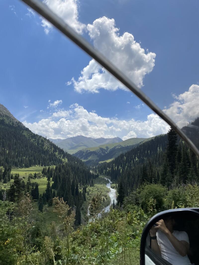 A scenic view of a mountain valley with a river flowing through it, captured from inside a vehicle, with the sky filled with fluffy white clouds.