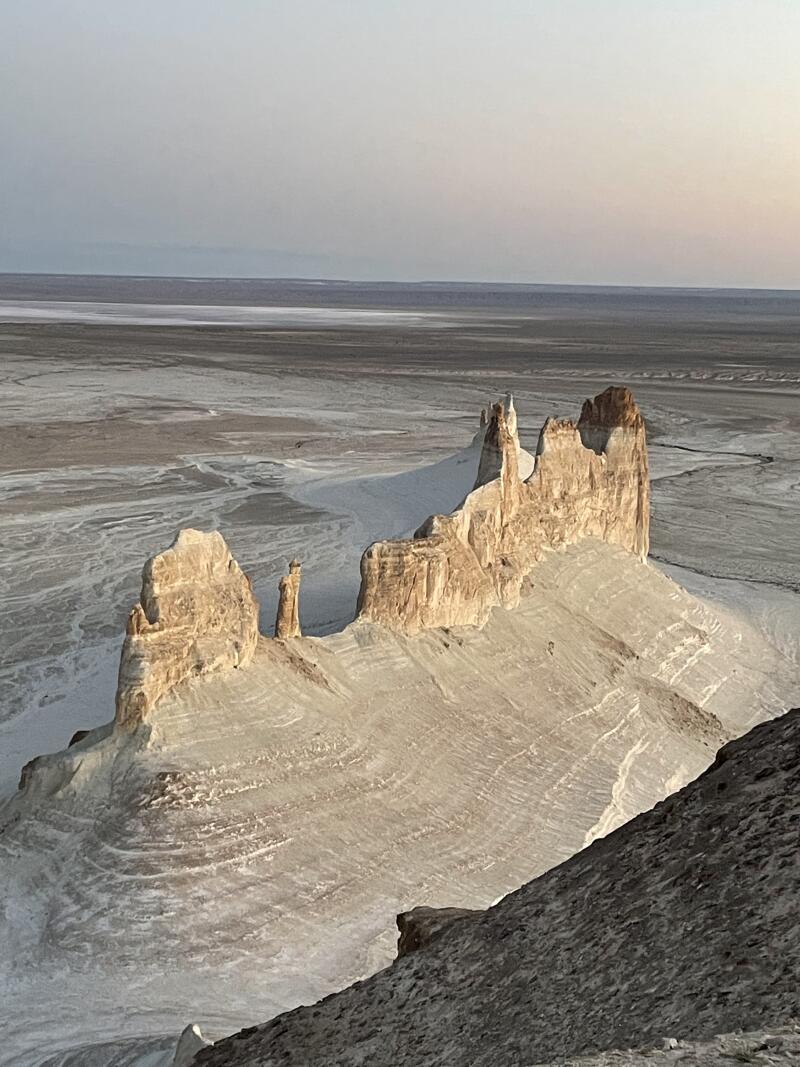 The image shows a landscape with unique rock formations in shades of white and brown, standing atop a layered, sandy hill, against a backdrop of a flat, expansive terrain under a pale sky.