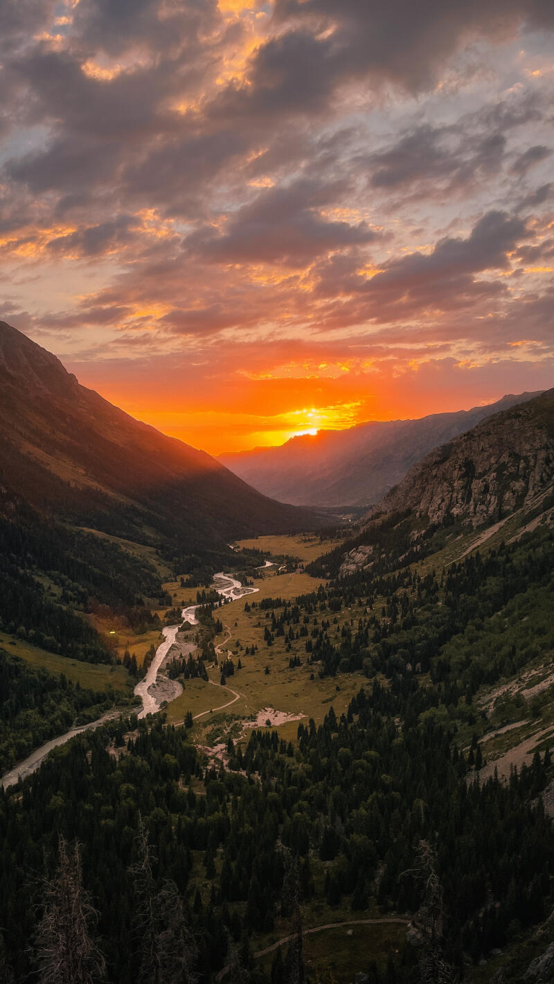 A scenic view of a valley with a river at sunset, featuring mountains, trees, and colorful clouds.