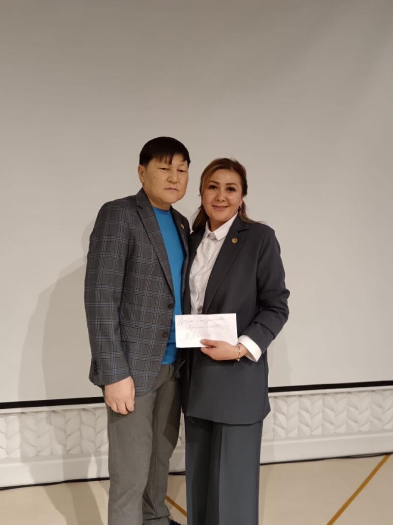 A man in a blue shirt and plaid blazer stands next to a woman in a dark suit holding a check that reads "Айса Сагумбаева, премия 91000".