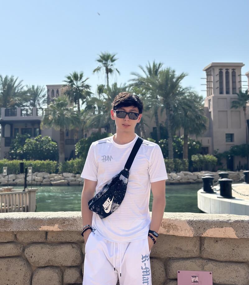 A person wearing sunglasses, a white t-shirt with 'RUN' printed on it, and white shorts with 'R-Mart' on the leg stands in front of a canal with palm trees and buildings in the background, also wearing a Nike fanny pack across the chest.