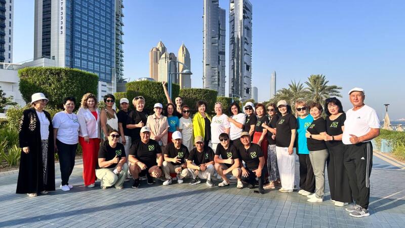 A group of people, mostly middle-aged and elderly, are posing for a photo in front of some skyscrapers and greenery; some are wearing black shirts with a green logo, while others have on casual attire.