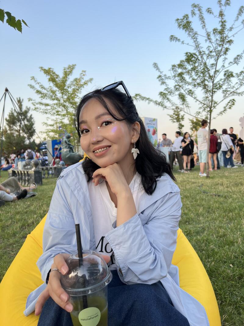 A smiling woman with glitter makeup and braces sits in a yellow beanbag chair, holding a drink with a straw; people and trees are visible in the background.