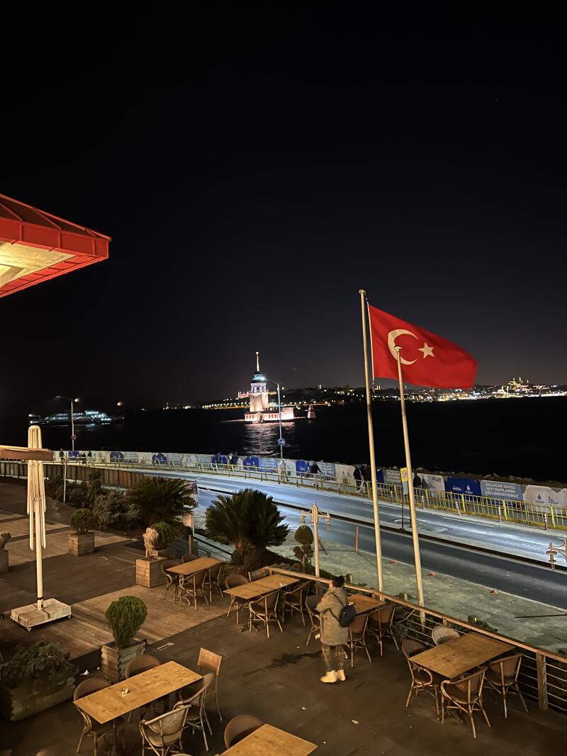 This is an outdoor restaurant scene at night with a Turkish flag waving in the foreground, overlooking the Maiden's Tower illuminated on the water.