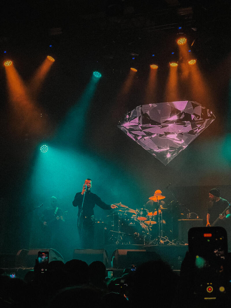 A band is performing on stage with a large diamond graphic in the background, illuminated by orange and green stage lights, and audience members are recording the performance with their phones.