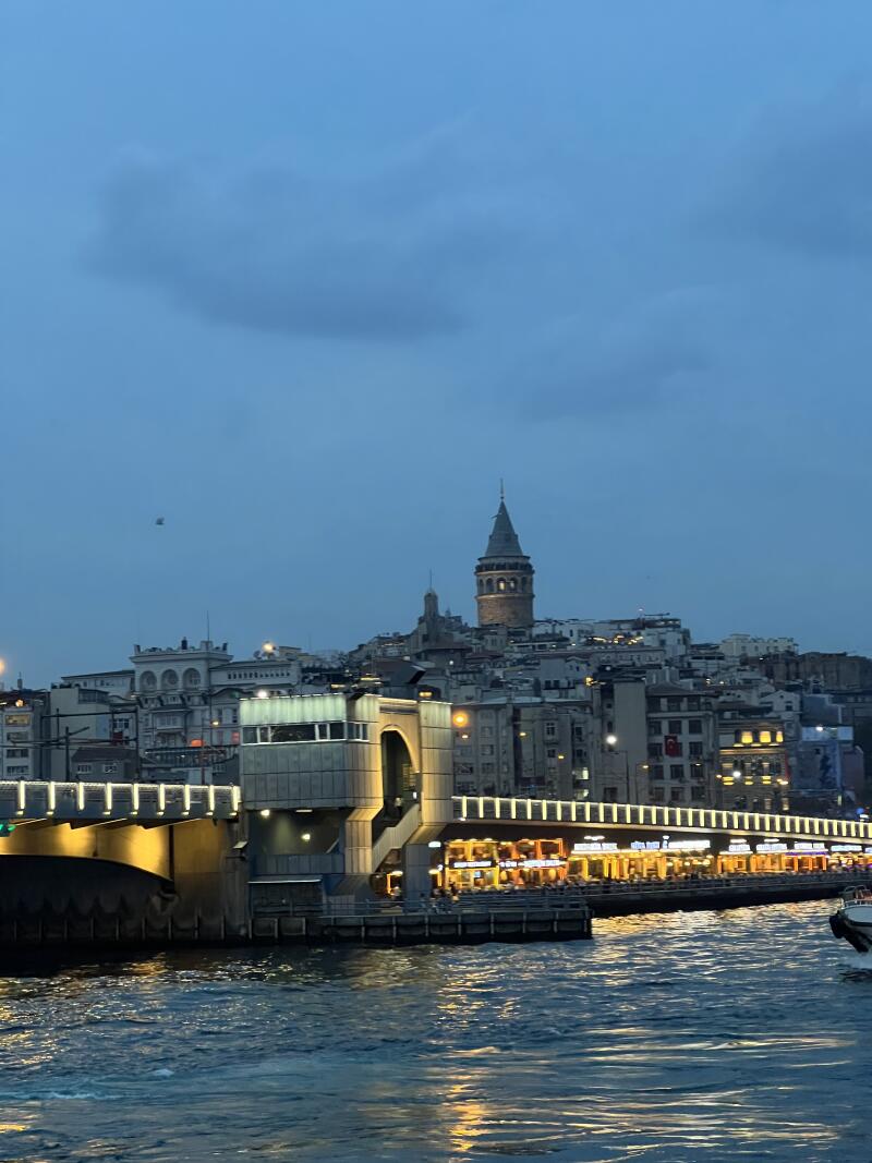 The photo shows a bridge with lights over the water, with buildings and the Galata Tower in the background under a cloudy sky.