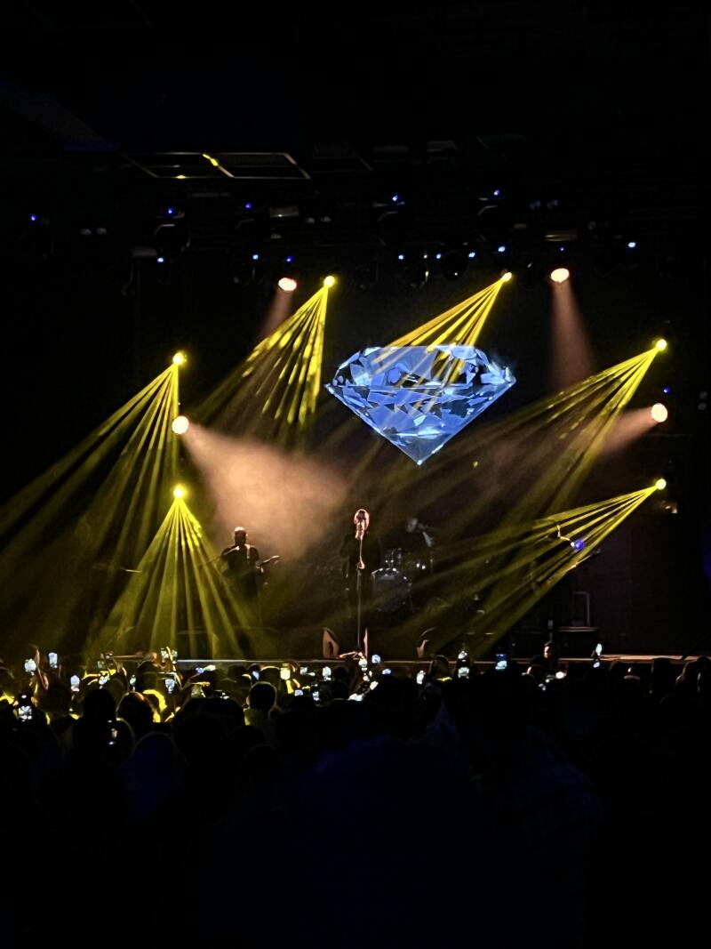 A band is performing on a stage with a large diamond graphic behind them, illuminated by yellow spotlights, while the audience records the performance with their phones.