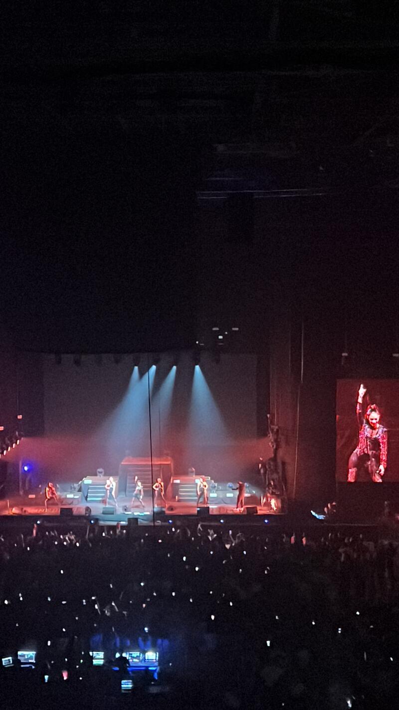 A crowd of people is watching a performance on a stage lit by spotlights, with a large screen displaying an image of a performer on the right side.