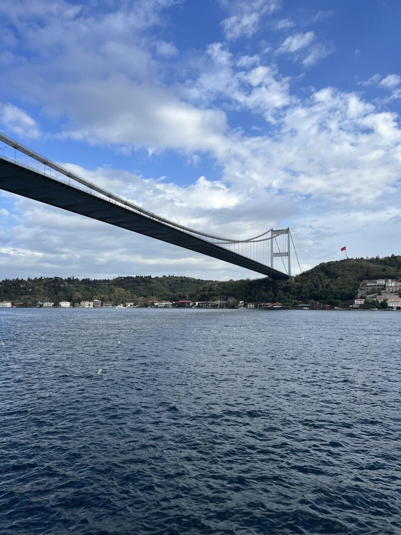 A bridge spans across a body of water under a blue sky with scattered clouds, with green hills and buildings visible in the background.