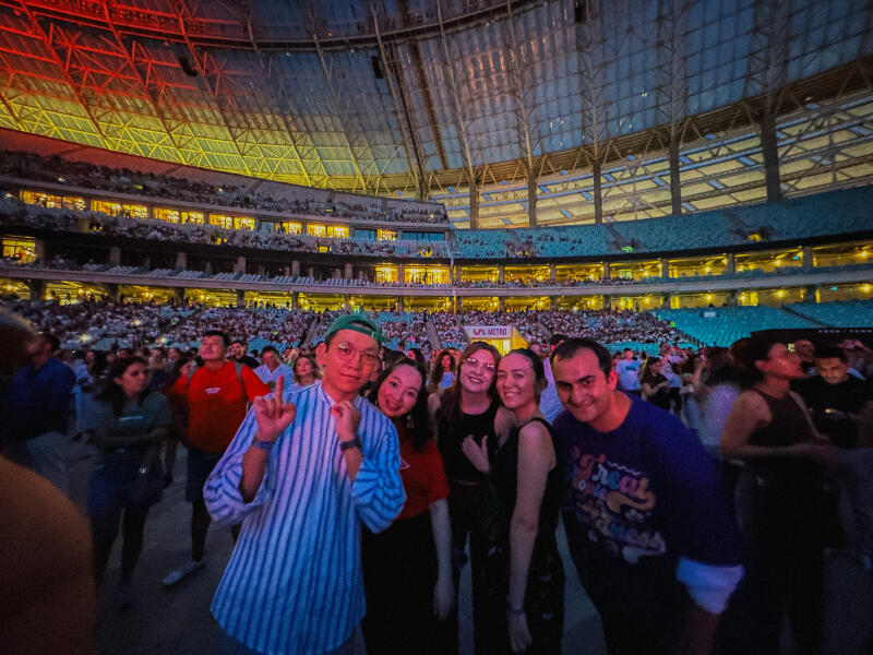 A group of people poses for a photo at a crowded indoor stadium with tiered seating, with a sign that reads 'NESTO' visible in the background.