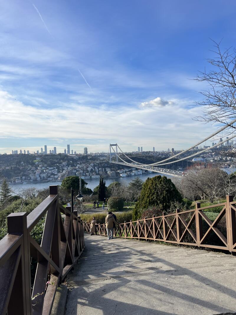 A view from a concrete path with a wooden fence overlooking a bridge spanning a body of water, with a city skyline in the background under a blue sky.