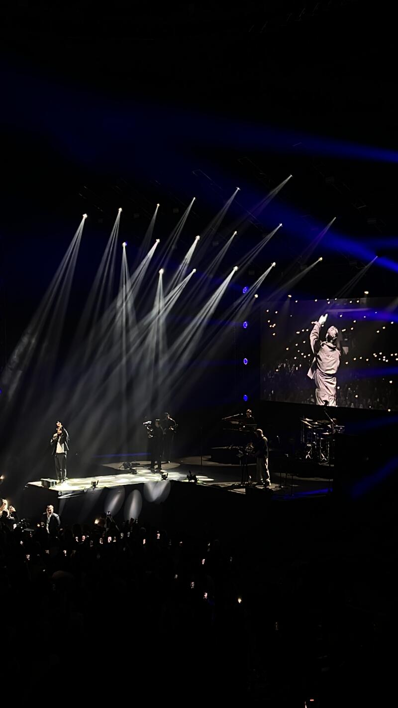 A band is performing on a stage with white spotlights shining on them, and a large screen behind them shows a person in a suit with their arm raised.