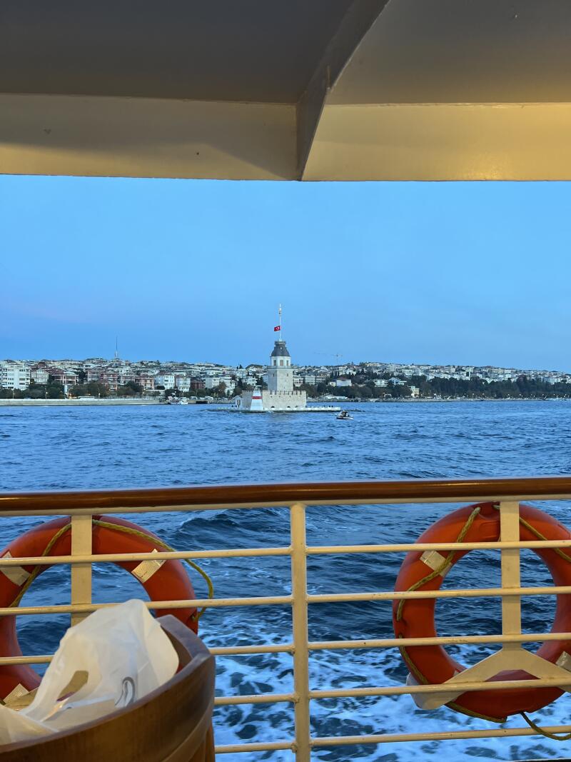 The photo shows the Maiden's Tower in Istanbul, Turkey, from the perspective of a boat with lifebuoys, with a cityscape in the background and a blue sky.