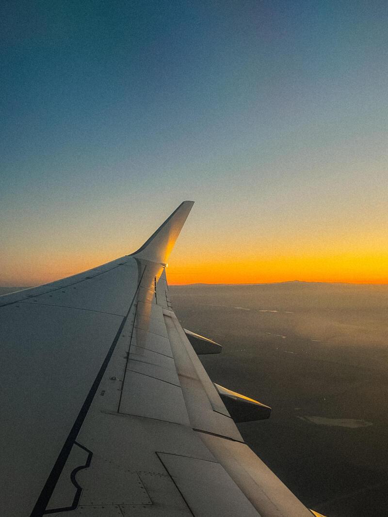 The view from an airplane window shows the wing and a colorful sunrise.
