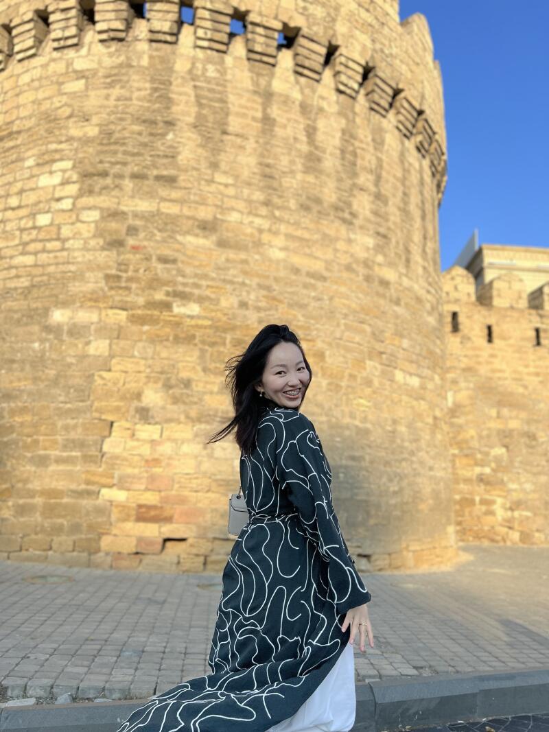 A woman in a black and white patterned coat smiles while standing in front of a stone fortress under a clear blue sky.