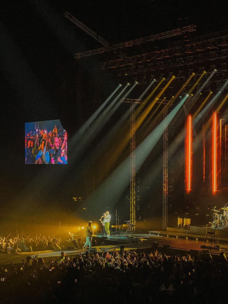 A concert stage is lit with spotlights and vertical orange lights, featuring a band performing in front of a large crowd, with a screen showing an image of the audience.