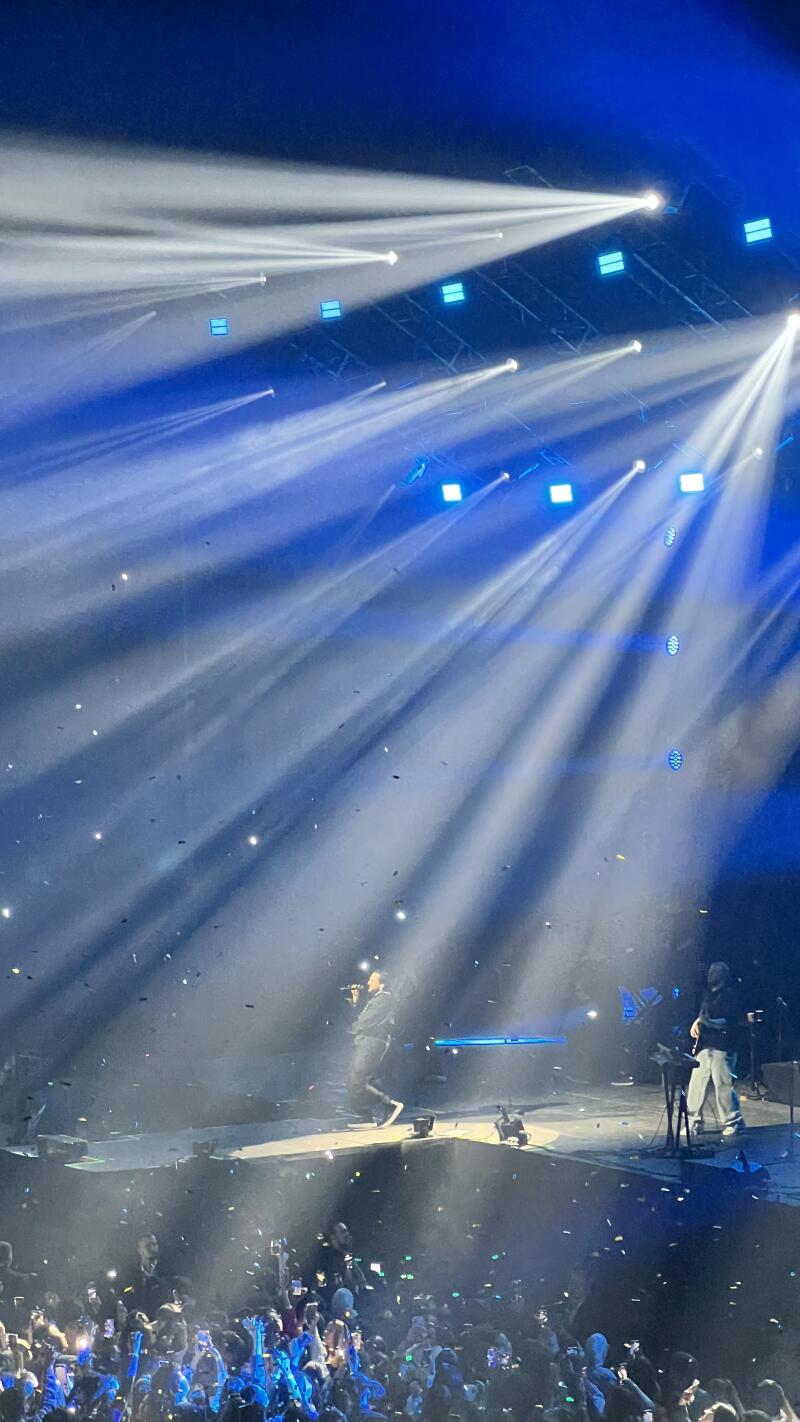 A crowded concert scene with a performer on stage under bright blue lighting and falling confetti, with audience members holding up phones.