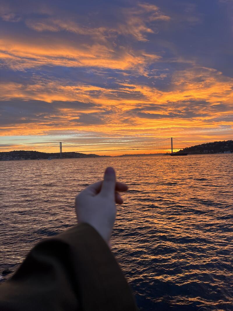 The photo captures a scenic view of a sea at sunset, with a bridge in the background and a hand making a heart shape in the foreground. The sky is filled with orange and blue hues.