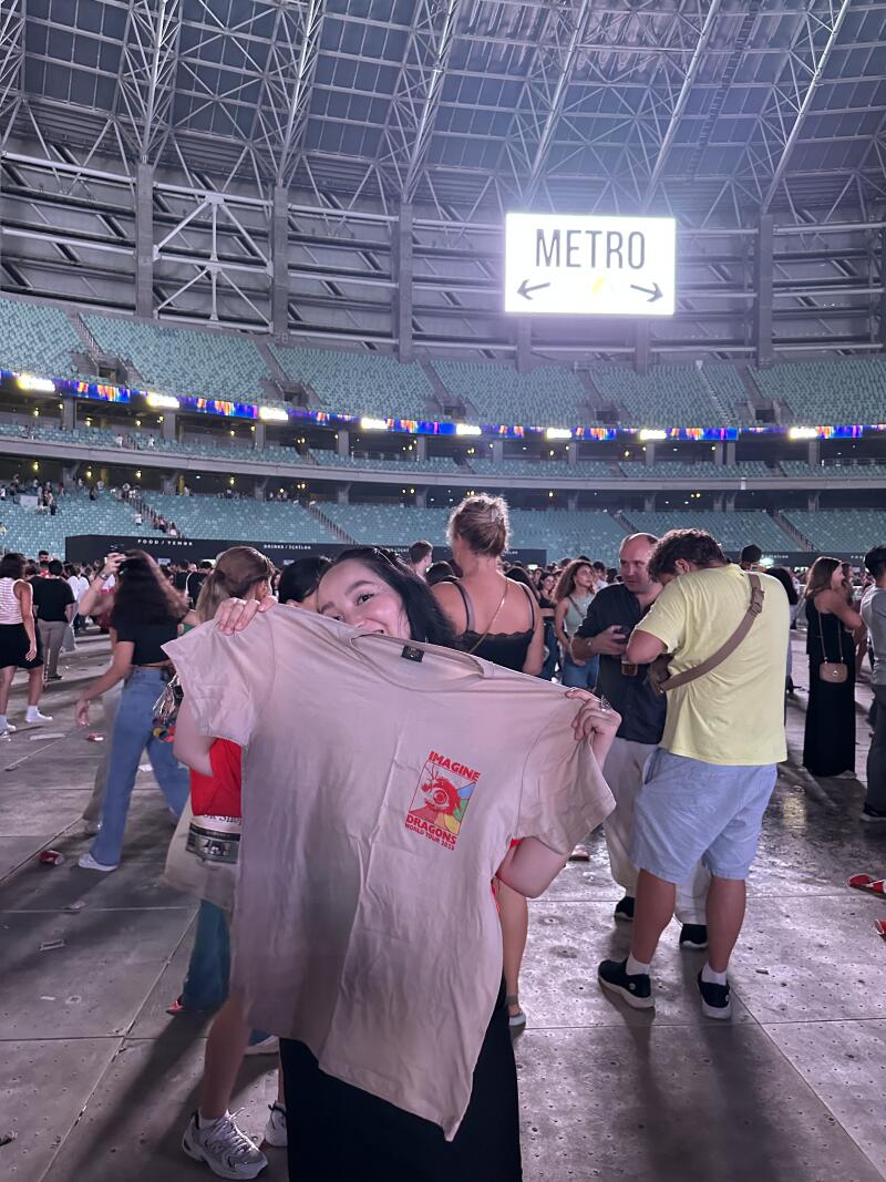 In a crowded stadium, a woman holds up a tan t-shirt with an "Imagine Dragons World Tour 2022" logo, while an overhead sign indicates "METRO" with directional arrows.