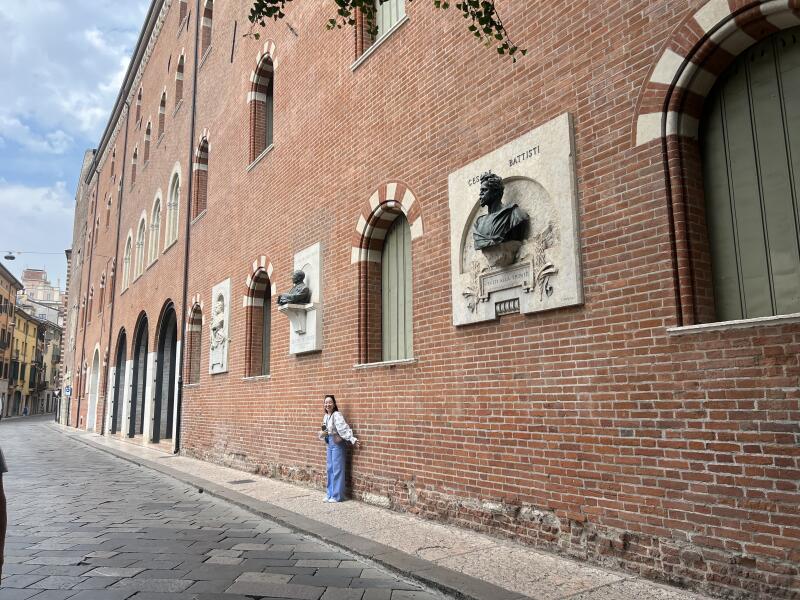 A woman stands against a brick building adorned with busts and plaques, positioned along a cobblestone street.