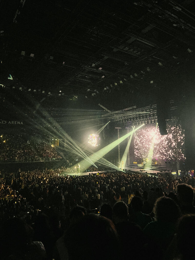 A concert hall filled with a large crowd is illuminated by stage lights and a large screen.