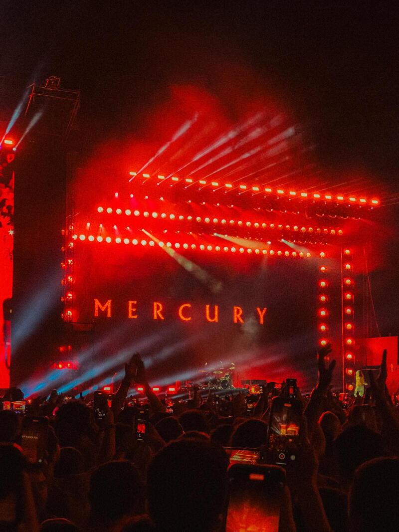 A crowd of people is silhouetted against a stage illuminated with red lights, with the word "MERCURY" prominently displayed on the backdrop.