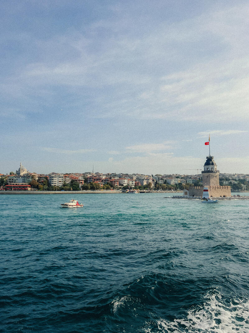 The image features the Maiden's Tower in Istanbul, Turkey, surrounded by the sea, with the coastline visible in the background and a small boat in the water.