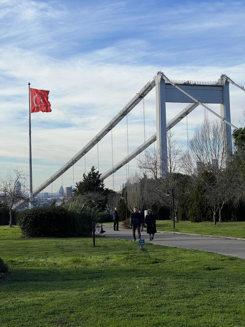 The photo shows a suspension bridge and a Turkish flag waving in the wind against a partly cloudy sky, with people walking on a path in a park-like setting.