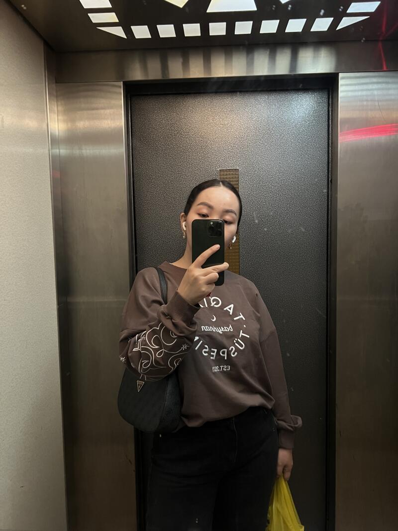 A woman is taking a selfie in an elevator, wearing a brown sweatshirt with white text and design on the sleeves, black jeans, and a black shoulder bag.
