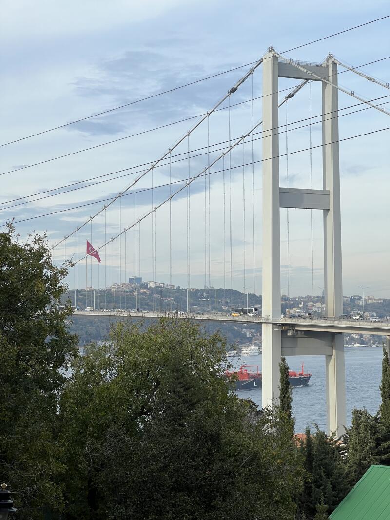 The image features a suspension bridge with a white tower and cables, with a Turkish flag waving in the background and a ship passing underneath.