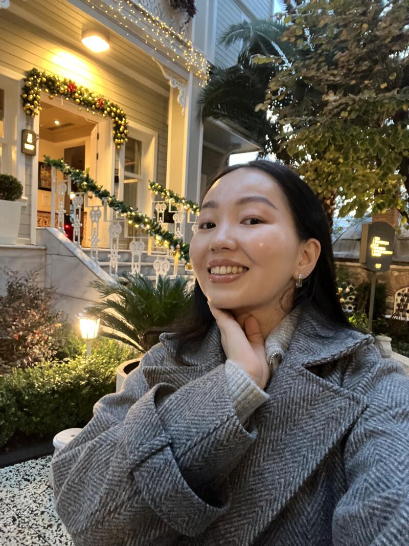 A smiling woman in a gray herringbone coat poses in front of a house decorated with festive lights and garlands.