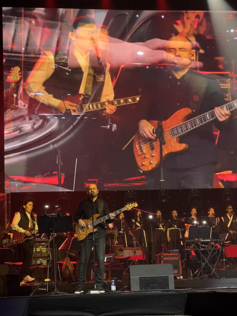 A stage with a band performing, featuring a guitarist and bassist in the foreground and a choir and keyboardist in the background, with a large screen above showing a close-up of the musicians.