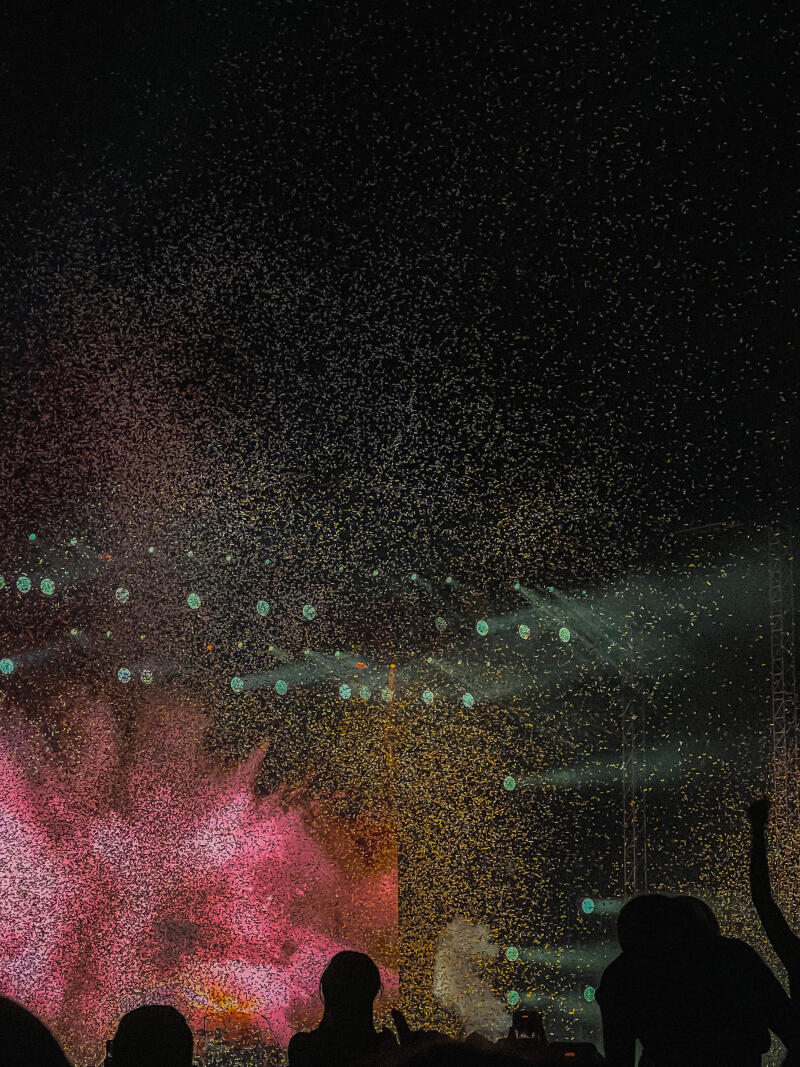 A crowd of people is silhouetted against a dark sky filled with confetti and stage lights.