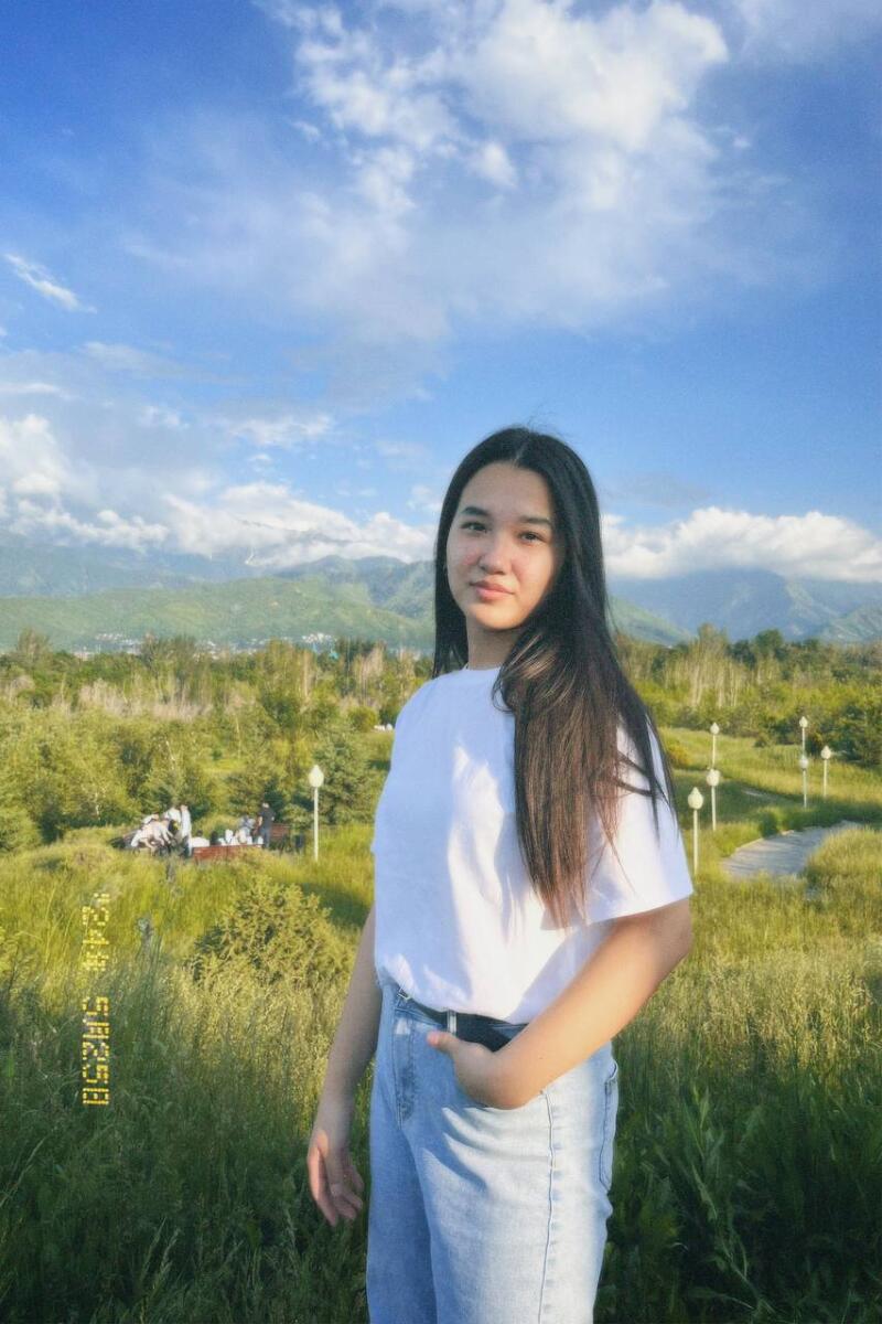 A young woman with long, dark hair stands in a field wearing a white t-shirt and jeans, with mountains and a blue, cloudy sky in the background.