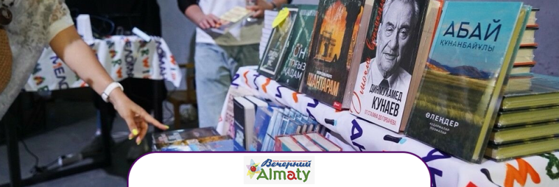 На столе выставлены книги, включая такие названия, как «Абай Кунанбайулы», «Динмухамед Кунаев» и «Шантарам», а рука женщины указывает на выбор; виден логотип «Вечерний Алматы».