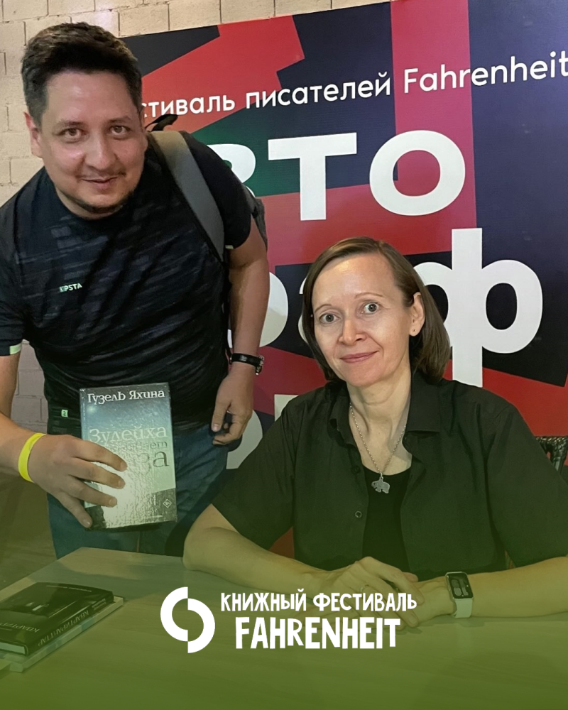 Мужчина держит книгу «Зулейха открывает глаза» Гузели Яхиной рядом с женщиной, сидящей за столом. Позади них баннер «Книжного фестиваля Fahrenheit», а логотип и название фестиваля также видны внизу изображения.