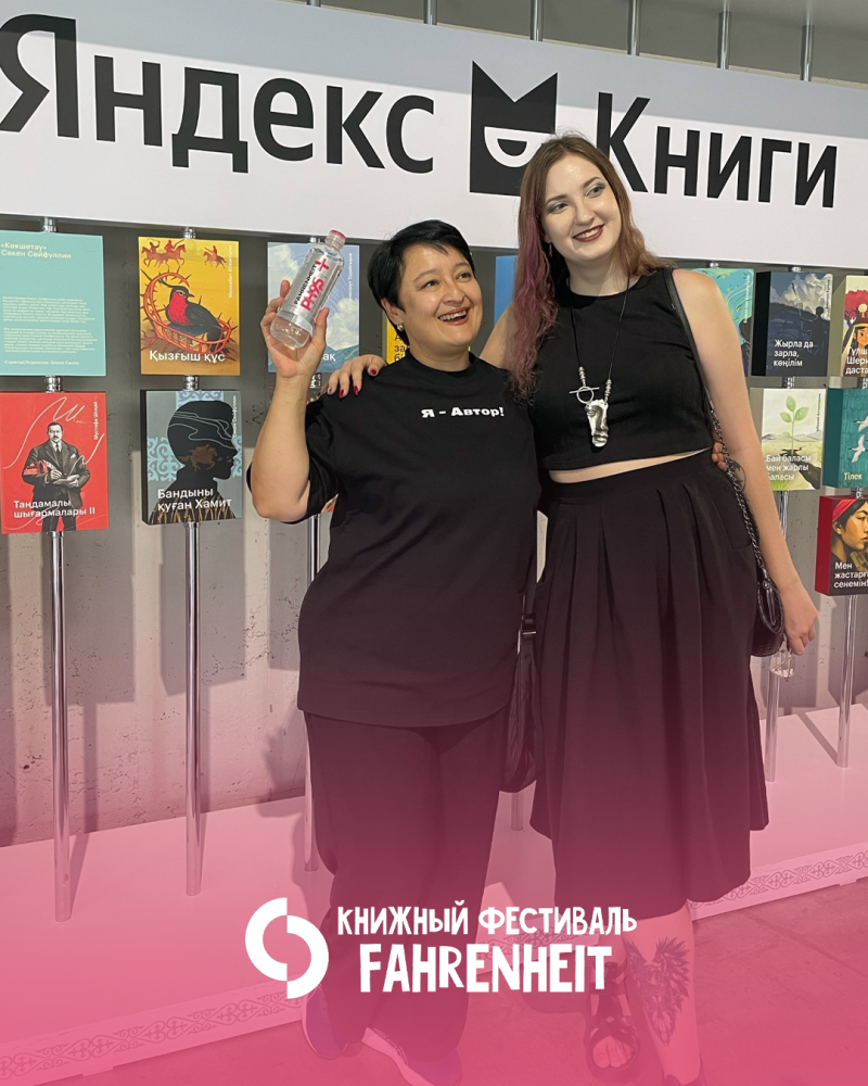 Две улыбающиеся женщины стоят перед вывеской «Яндекс Книги» и книжной витриной. Одна женщина держит бутылку воды и одета в футболку с надписью «Я - Автор!», в то время как другая одета в черный укороченный топ и юбку. Внизу изображения находится наложение текста «КНИЖНЫЙ ФЕСТИВАЛЬ FAHRENHEIT».