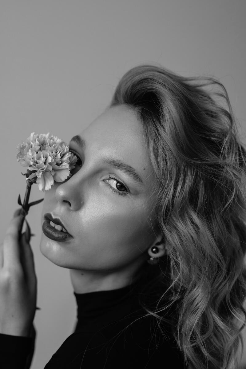 A grayscale portrait of a woman with wavy hair, wearing a black turtleneck, and holding a flower in front of her eye.
