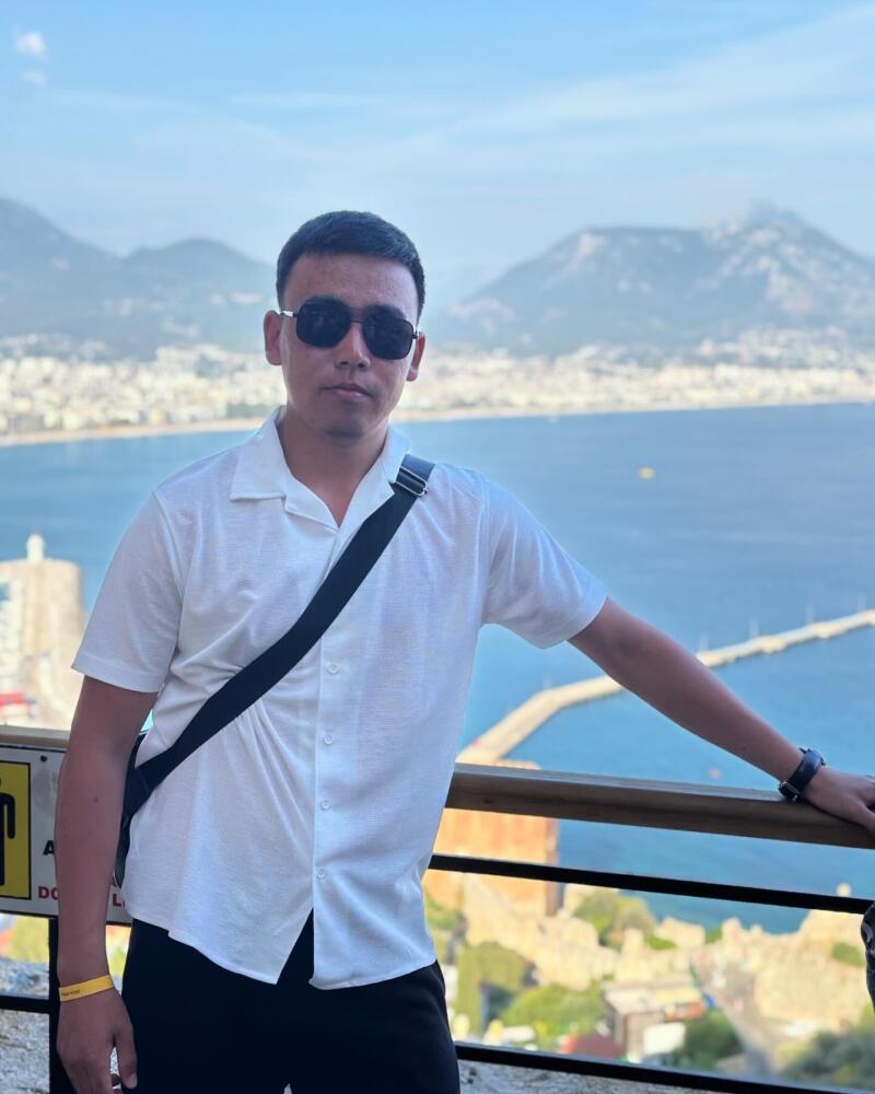 A man in sunglasses and a white shirt stands in front of a scenic view of the ocean and mountains.