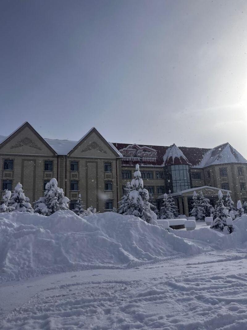 The photo shows the 'Altai Village Mountain Resort' building, surrounded by snow-covered trees and a thick layer of snow on the ground.