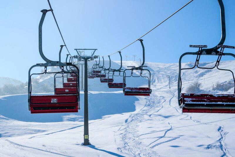 The photo shows a ski lift with red seats extending up a snow-covered mountain under a clear blue sky, with the number '104' visible on one of the seats.